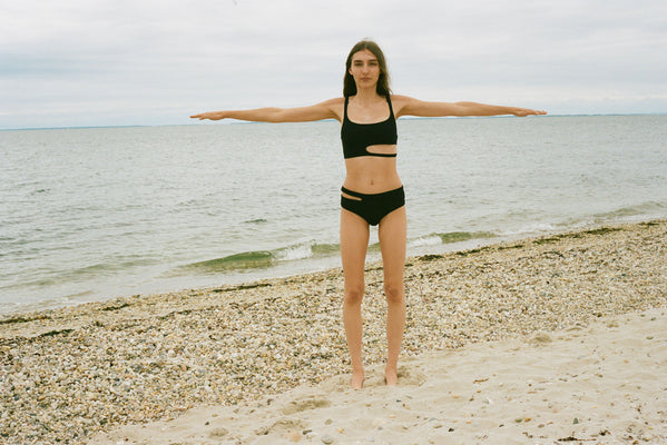 A woman in a black bikini standing on a beach with her arms out.