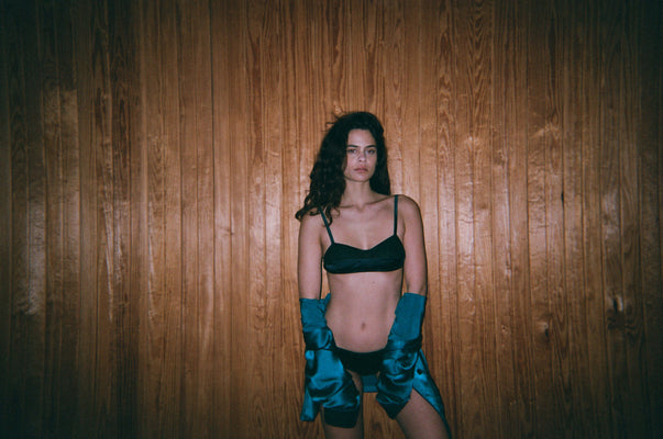 Woman in black underwear and a bra with blue gloves against a wooden wall.