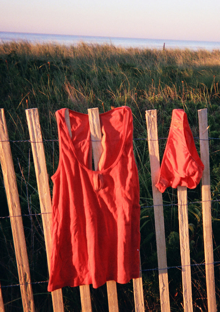 Orange tank top and matching swim bottoms hung on fence next to beach.