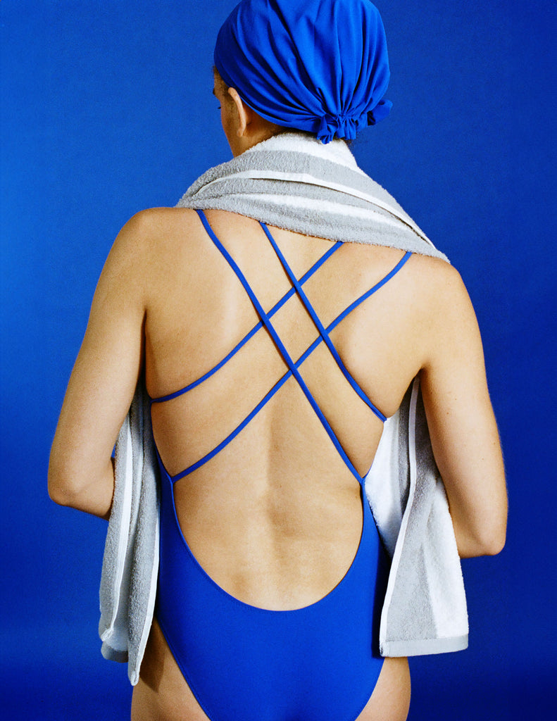 Person wearing an Araks blue swimsuit with criss-cross straps with a swim aht against a blue background
