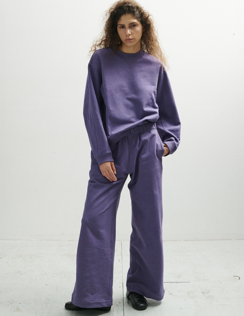 A model posing in the Araks Vivienne crewneck and the matching purple Vero sweatpants in the shade Violette, while standing against a plain off-white studio background.