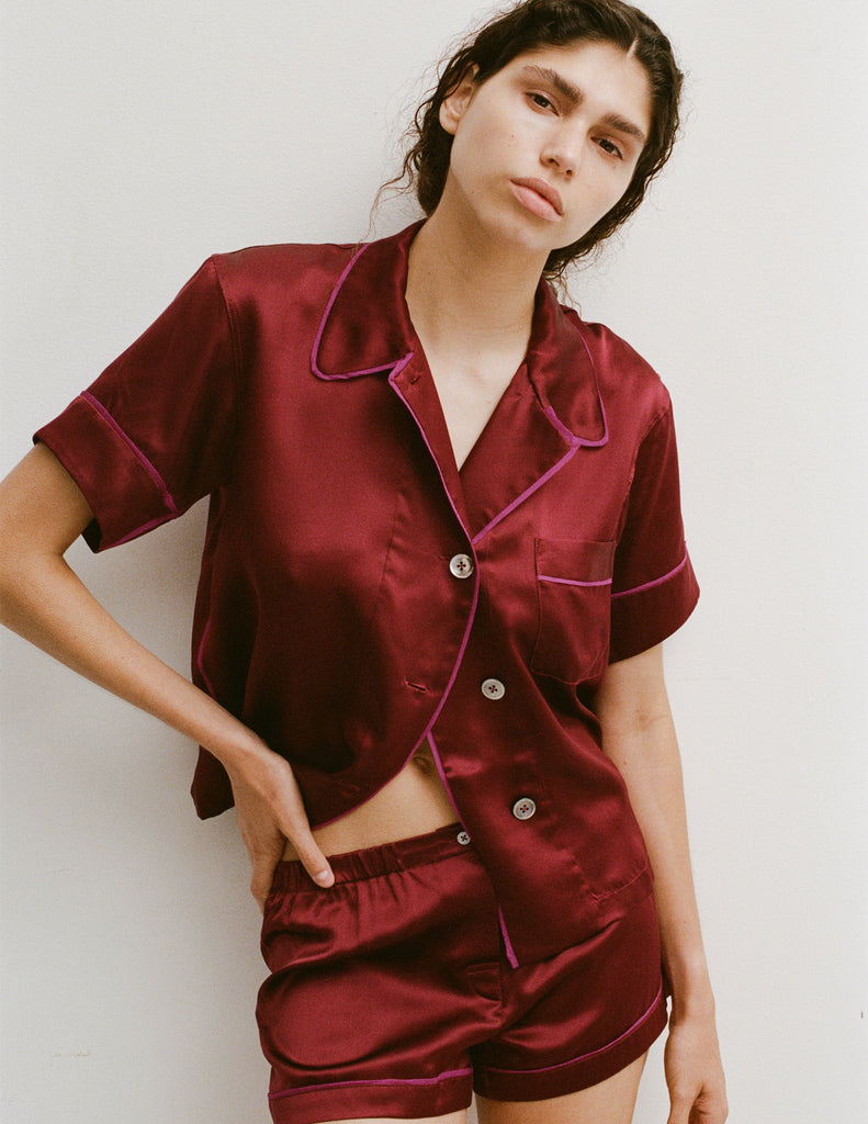A woman modeling the Araks Shelby silk pajama top with the Tia pajama shorts in Garnet against a light gray studio background