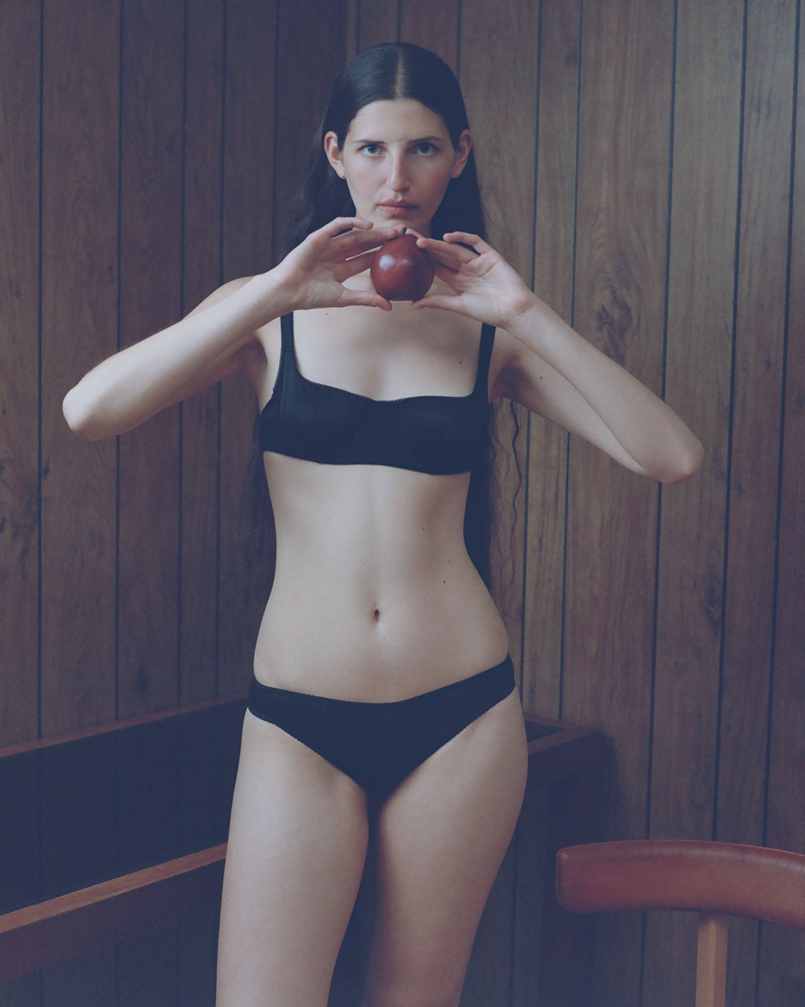 A model standing in front of a wooden wall holding an apple wearing the araks silk penelope bralette and gwyneth panty in black