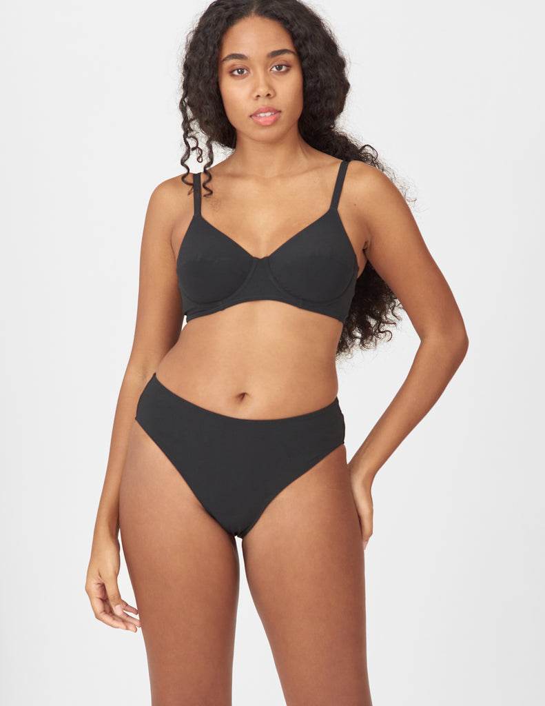A woman modeling a two-piece black bikini set against a plain light gray studio background.