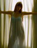 A model posing in sheer curtains while wearing a semi sheer Araks Raffaelle Slip. 