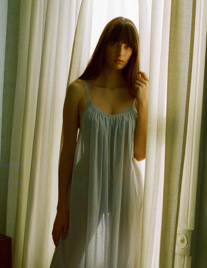 A model posing in sheer curtains while wearing a semi sheer Araks Raffaelle Slip. 