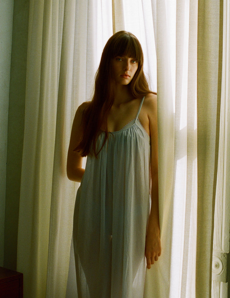 A model posing in sheer curtains while wearing a semi sheer Araks Raffaelle Slip. 