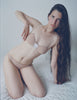 A model sitting on a bed in the araks cotton crepe and silk uma bralette and harriet panty in brulee brown and lavender
