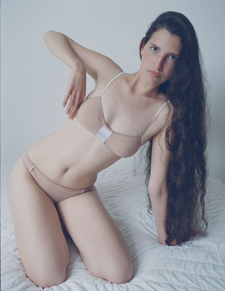 A model sitting on a bed in the araks cotton crepe and silk uma bralette and harriet panty in brulee brown and lavender