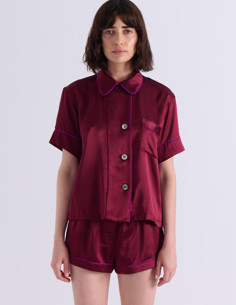A woman modeling the Araks Shelby silk pajama top with the Tia pajama shorts in Garnet against a light gray studio background