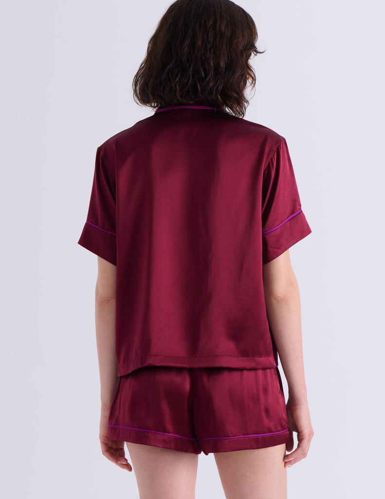 A woman modeling the Araks Shelby silk pajama top with the Tia pajama shorts in Garnet against a light gray studio background