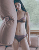 A model in the silk georgette and silk charmeuse beatrice bralette and aaron panty in noisette brown and purple standing in front of a couch holding a pillow