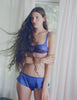 A model in the araks silk gita underwire bra and gal hipster in tanzanite blue standing in front of a door with curtains