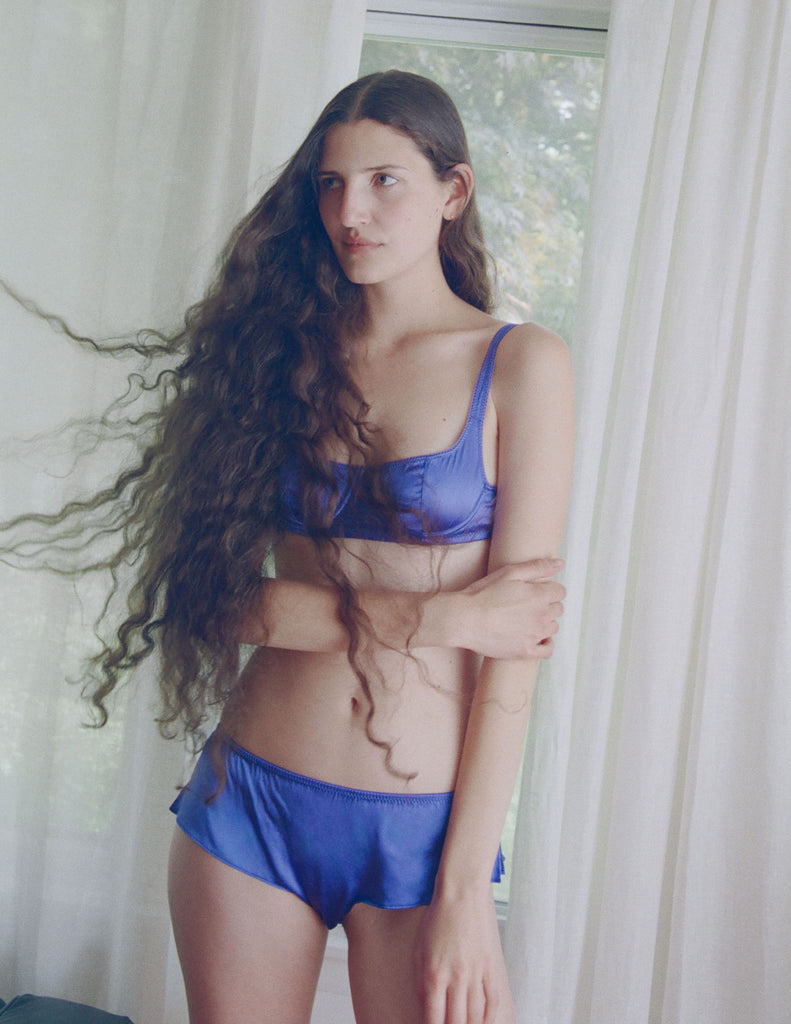 A model in the araks silk gita underwire bra and gal hipster in tanzanite blue standing in front of a door with curtains