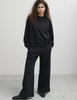 A model wearing the Araks Valerie black crewneck sweatshirt and matching black wide-leg Araks Vero sweatpants, standing against a light gray studio wall.