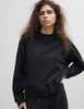 A model wearing the Araks Valerie black crewneck sweatshirt standing against a light gray studio wall.