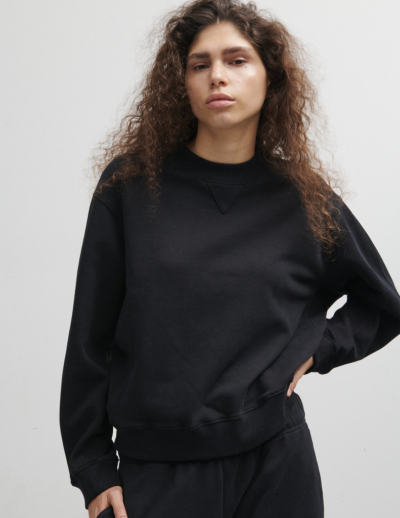 A model wearing the Araks Valerie black crewneck sweatshirt standing against a light gray studio wall.