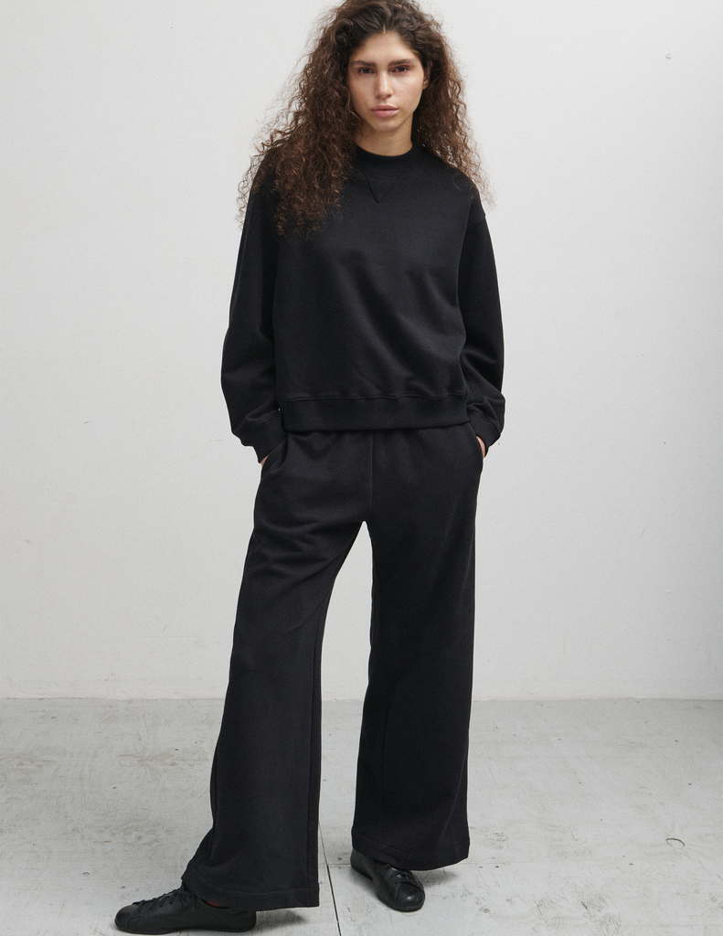 A model wearing the Araks Valerie black crewneck sweatshirt and matching black Vero sweatpants, standing against a light gray studio wall.