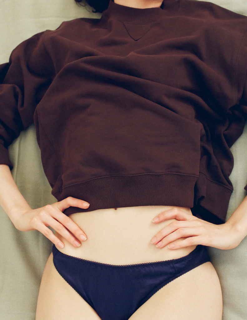 A close-up of a model lying down, wearing the Araks Valerie sweatshirt in the dark brown shade Mill and navy blue underwear.