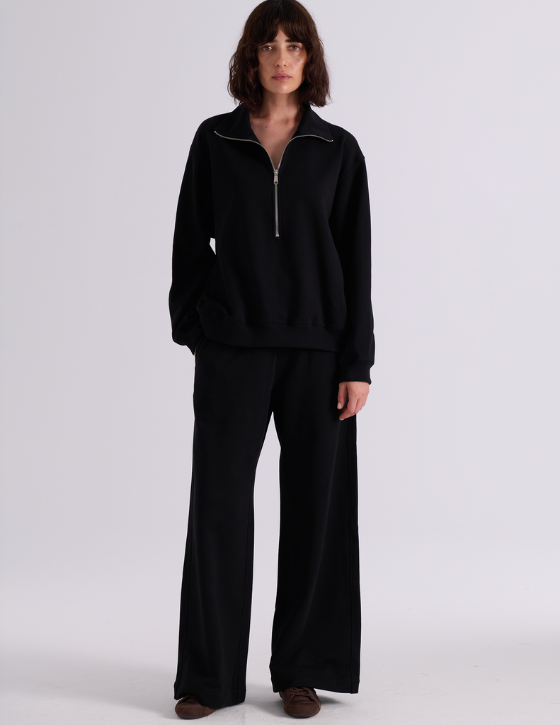 A model wearing the Araks Vivienne black half-zip sweatshirt and matching Vero wide-leg sweatpants, standing against a light gray studio background.