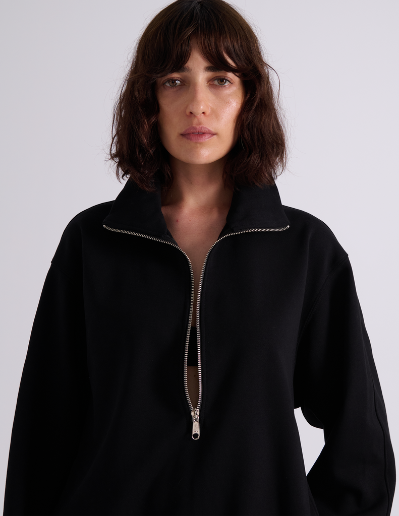 A model wearing the Araks Vivienne black half-zip sweatshirt, standing against a light gray studio background.