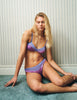 Woman in a Araks Beatrice Bralette in purple  sitting on the floor against a textured wall.
