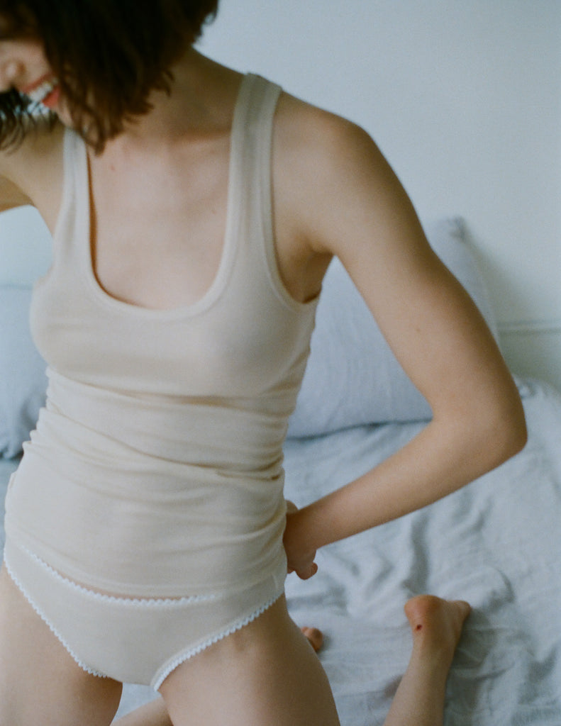 A model on a white bed wearing an off white James tank in the shade undyed. 