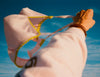 Person holding a pink bikini top with yellow trim against a blue sky.