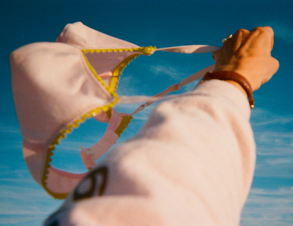 Person holding a pink bikini top with yellow trim against a blue sky.