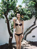 Person in a maroon ovsanna bikini top standing under a fruit tree with a white wall background
