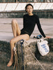 Model wearing the Araks Undine Rashguard in Black, a long-sleeve fitted rash guard with a crew neck, paired with black bikini bottoms, sitting on a pier