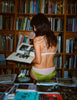 Woman in lingerie set reading a book in a room filled with books and magazines.