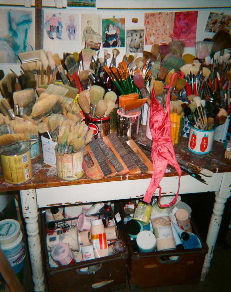 Art supplies including brushes, paint cans, and a pink bra on a cluttered desk. 