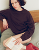 A model sitting on a bed, wearing the Araks Valerie crewneck sweatshirt in the dark brown shade Mill. 