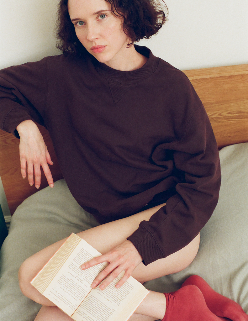 A model sitting on a bed, wearing the Araks Valerie crewneck sweatshirt in the dark brown shade Mill. 