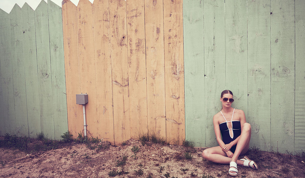 Woman in front of fence, wearing navy one piece with white piping and halter neck detail, and white shoes and sunglasses.