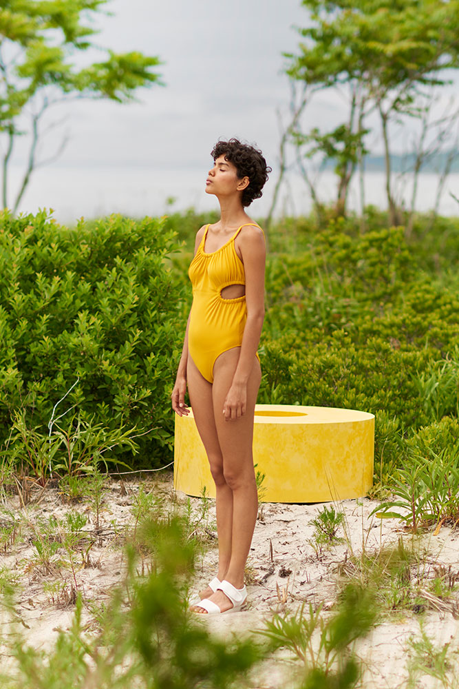Woman outside wearing a yellow one piece swimsuit with ruched neckline, side cut outs and a tie in back.