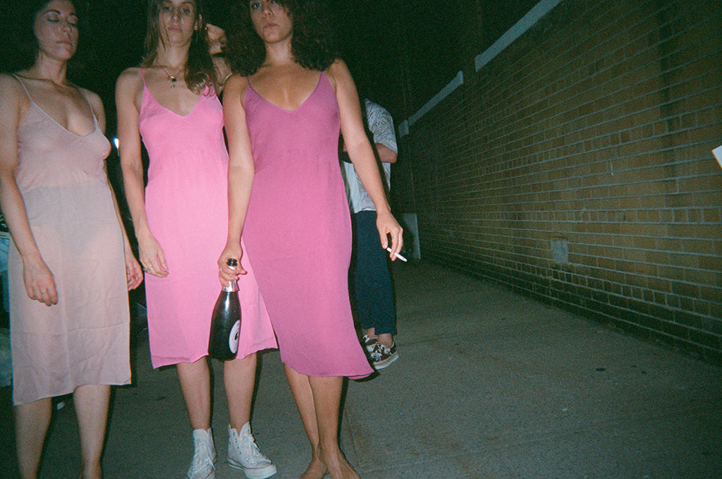 Three women outside, one in a pale pink, second light pink, and third in pink wearing slips