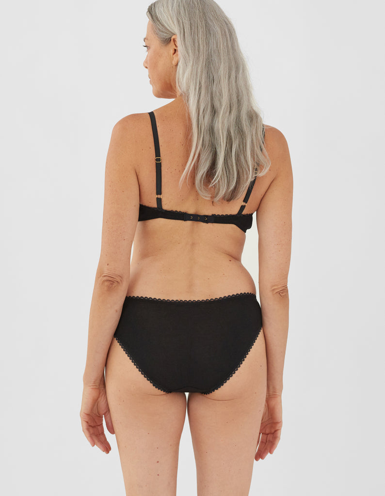 Back view of woman wearing araks organic cotton black isabella panty with white trim, and matching bralette.