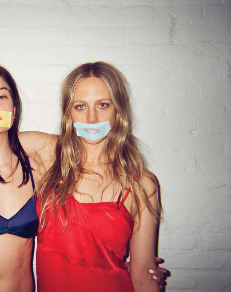 Two women with their arms around each other, with colored pieces of tape covering their mouths. Wearing red silk top and blue bra. 
