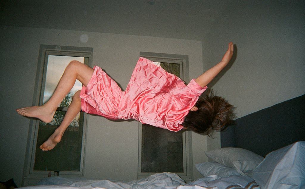 Woman jumping on a bed wearing pink silk collared short-sleeved sleep shirt with left breast pocket and contrast piping and matching shorts.