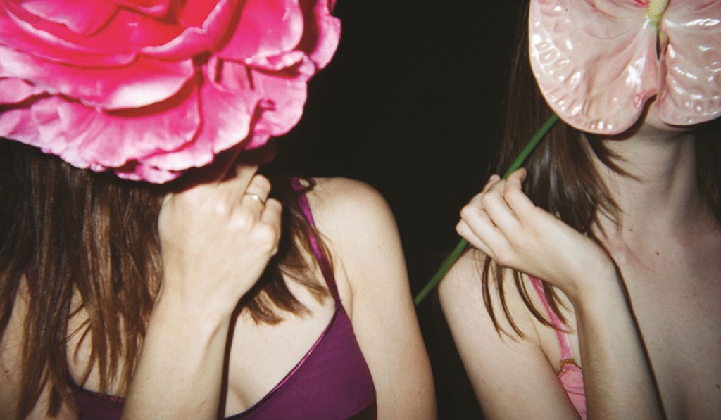 Two women holding flowers in front of their faces wearing burgundy demi wireless bralette with burgundy silk charmeuse, and burgundy shiny straps. Other woman wearing pink bralette with orange scallop trim. 