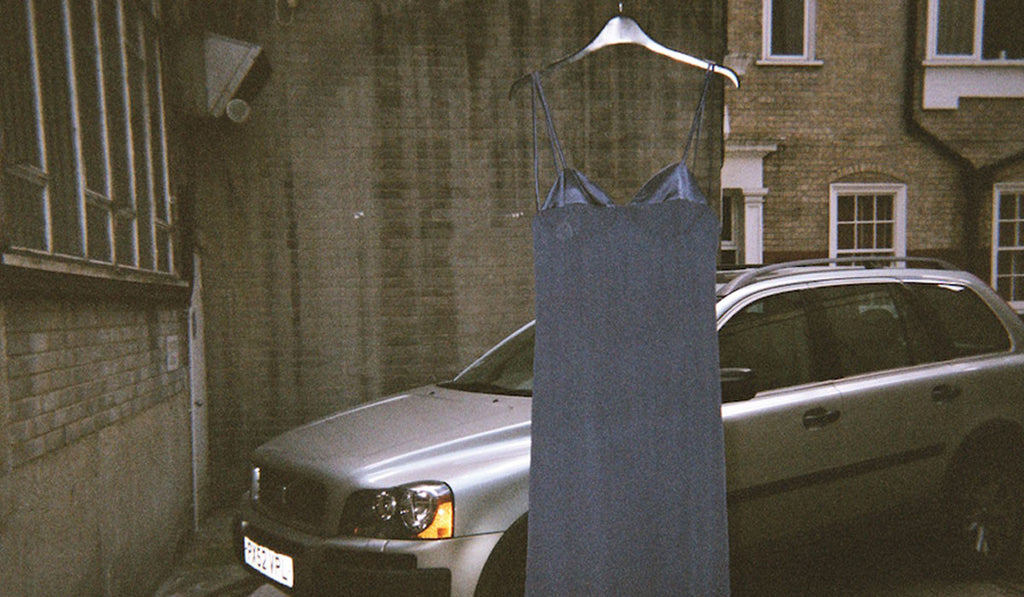 Blue silk charmeuse slip dress hung on a hanger in front of a car and building. 