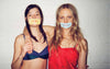 Two woman with tape over their mouths, one is wearing blue bralette with silk charmeuse detailing. Other woman wearing red silk charmeuse slip dress.
