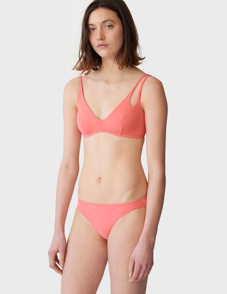 Woman wearing peach low-rise swim bottoms with matching bikini top with asymmetrical crisscross strap.