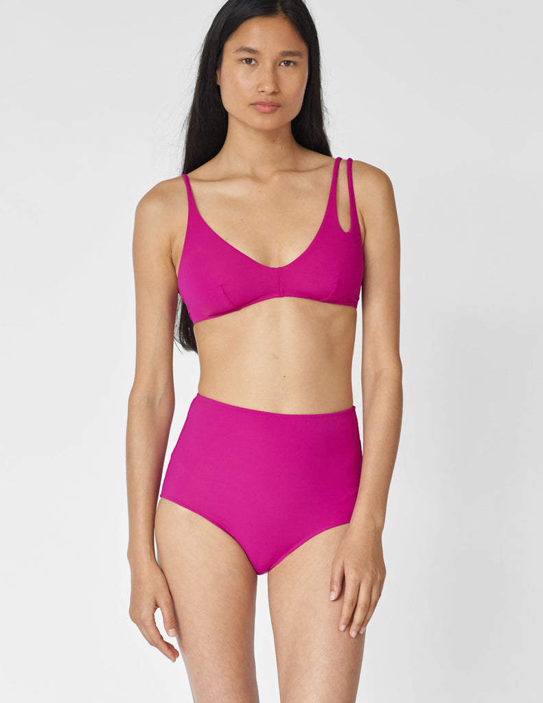 Woman wearing a hot pink bikini top with asymmetrical crisscross strap with matching high-waisted swim bottoms.