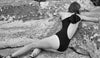 Back shot of woman sitting on rocks wearing black one-piece with single shoulder maillot and wide ties in a bow at back. 
