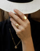 White woman grabs the rim of her panama hat and shows four solid 18k yellow gold rings. She wears a solid 18k yellow gold Dutch Coin ring. Large circle of gold with a slight puff in the center and softened, thick edges on her index finger.